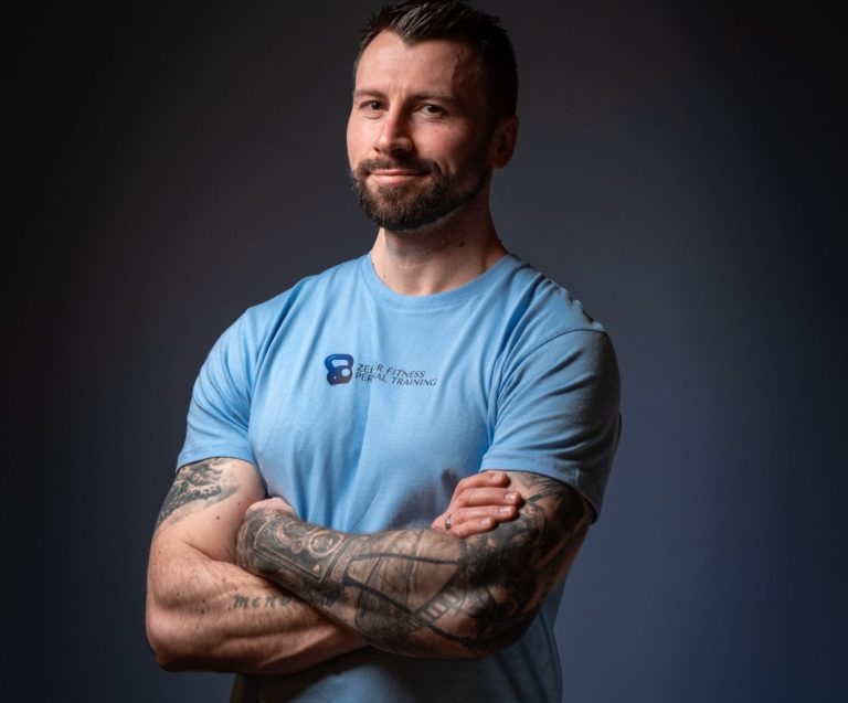 Coach Patrick Ein lächelnder Mann mit Tattoos, in einem blauen T-Shirt und verschränkten Armen.
